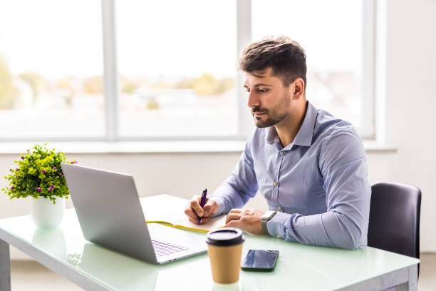 istock Entrepreneur writing notes sitting at his desk. Young man making business plans with papers and computers on his desk in office. 1360337863