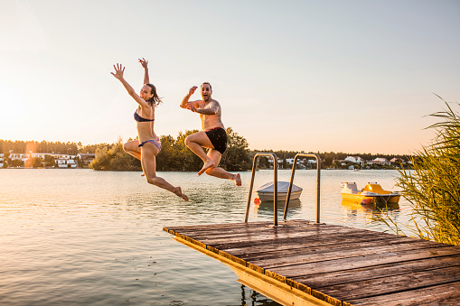 Two friends jumping off pier in the lake together at beautiful orange sunset