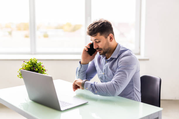istock Confident young man working on laptop and talking on the mobile phone while sitting at his working place in office 1360338859