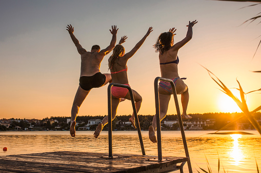 Three friends jumping off pier in the lake together at beautiful orange sunset