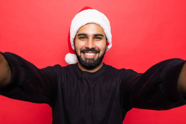 istock Handsome young man wearing santa hat, taking selfie on mobile phone , standing over red background 1360344903