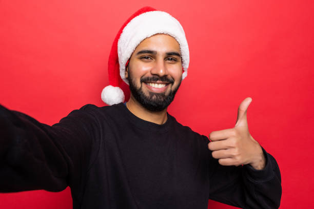 istock Handsome young man wearing santa hat, taking selfie on mobile phone , standing over red background 1360345029