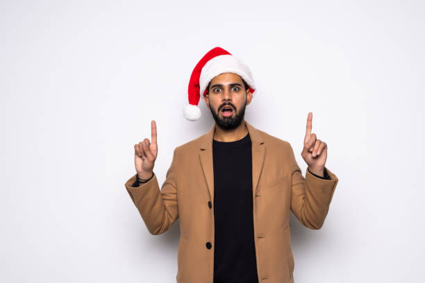 istock Young handsome man wearing a Santa hat over white background amazed and surprised looking at the camera and pointing up with fingers 1360349068