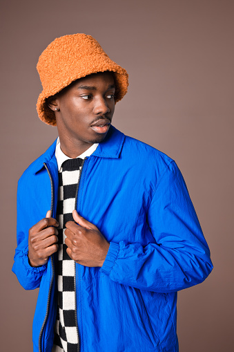 Portrait of fashionable young man wearing checker chess vest, blue jacket and orange bucket hat looking away. Studio shot on brown background.