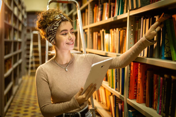 istock Young librarian picking book from bookshelf and holding tablet 1360367271