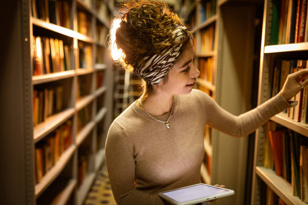 istock Young librarian picking book from bookshelf and holding tablet 1360367377