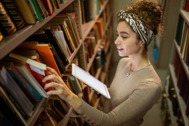 istock Young student holding tablet and searching book at library 1360367465