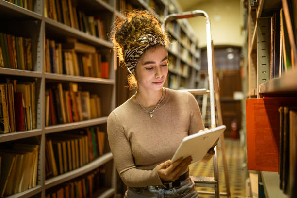istock Young female student at library trying to find book while using tablet 1360367629