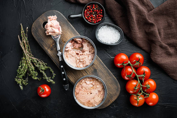 canned wild yellowfin tuna, on wooden cutting board, on black background with herbs and ingredients, top view flat lay - tuna imagens e fotografias de stock