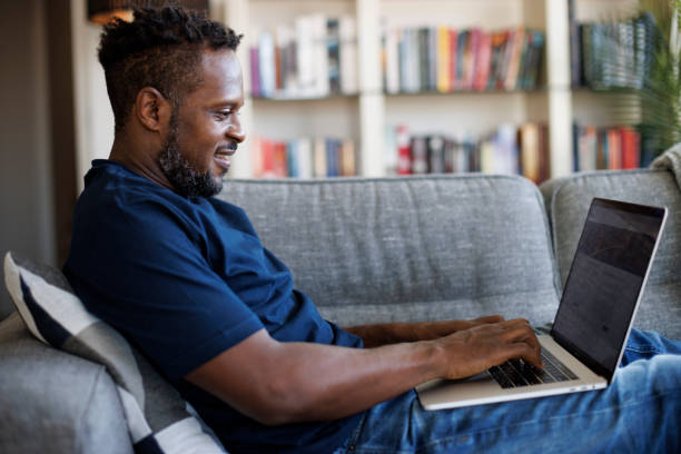 relaxed smiling man sitting on sofa and using laptop - applying imagens e fotografias de stock