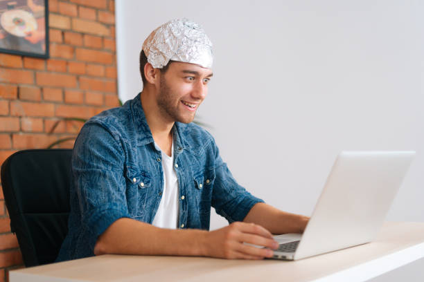 Medium shot of crazy male conspiracy theorist wearing aluminum foil protect brain watching shocked online video content using laptop computer. Side view of crazy smiling male conspiracy theorist wearing aluminum foil protect brain watching shocked online video content using laptop computer, sitting at desk in living room with modern interior tin foil hat stock pictures, royalty-free photos & images