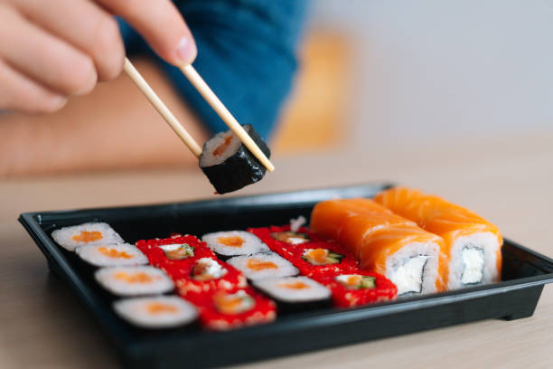 istock Close-up front view hands of unrecognizable young man holding sushi with chopsticks sitting at desk. 1360379189