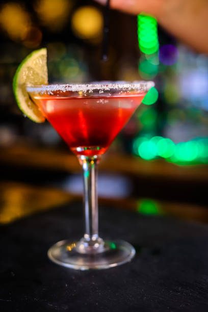 istock In a funnel-shaped glass on a long stem, a two-tone cocktail, dark red below, translucent white above, a lime slice on the edge of the glass. Filming of the bar menu against the background of bright lights in bokeh. 1360379887