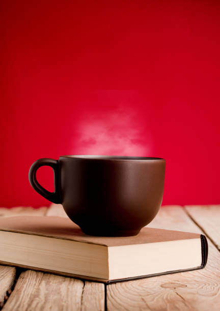 brown cup of coffee with hot steam on book on rustic wooden table, red background