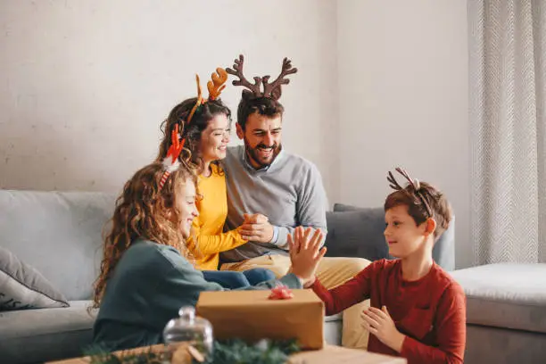 A four-member family is in the living room. They are enjoying during New Year's holiday. Kids are on the floor, sitting and playing. A young couple is watching them from the sofa bed. It is Christmas time and New Year and everybody have antlers on their heads.