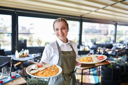 Beautiful Smiling Blond Waitress Bringing Two Plates Of Scrambled Eggs Breakfast To Restaurant Table