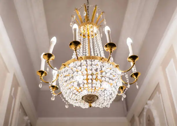 Beautiful chandelier hanging from ceiling. Classic gold plated design with glass crystals. Bright lights viewed from below with classy glass expensive bulbs.