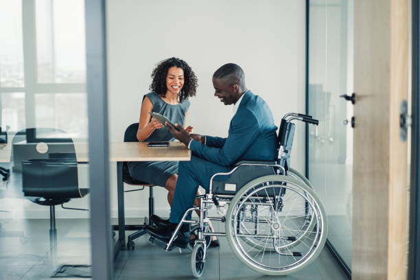 Business partners in meeting. Shot of an african-american businessman in wheelchair having discussion with his female colleague in the board room. Photo of elegant businesswoman working with her disabled colleague in the office. Corporate business persons discussing new project and sharing ideas in the workplace. wheelchair stock pictures, royalty-free photos & images