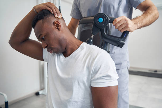 Tranquil patient undergoing percussive therapy performed by physiatrist Calm young African American man having his neck massaged with percussion gun percussion instrument stock pictures, royalty-free photos & images