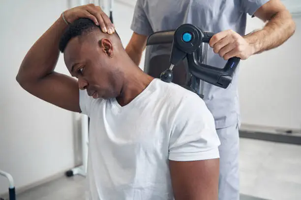 Calm young African American man having his neck massaged with percussion gun