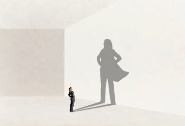 A woman stands with her hands on her hips as she looks up at her shadow that is cast on the wall in front of her that shows her wearing a cape and expressing  power and confidence.