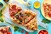 Image of four pizza slices with various toppings including chicken and sweetcorn, pepperoni, Pizza Margherita and vegetable with black olives chopping board with greaseproof paper, salt cellar, tomatoes, blue and yellow split background, elevated view