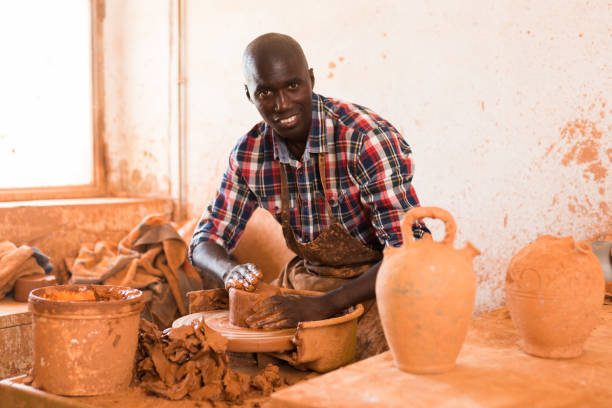 Smiling guy working with clay on potter wheel Smiling African American guy enjoying work with clay on potter wheel in ceramics workshop hobbyist stock pictures, royalty-free photos & images