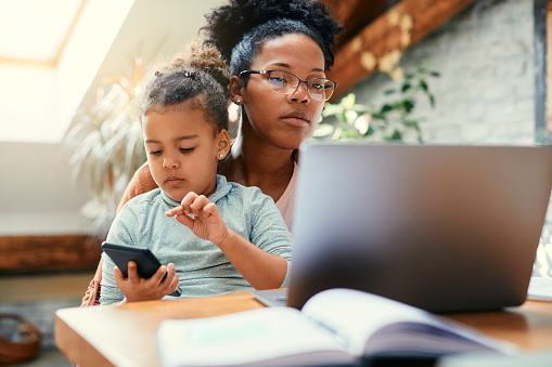 Niña negra usando un teléfono inteligente mientras su madre está trabajando en una computadora en casa. photo