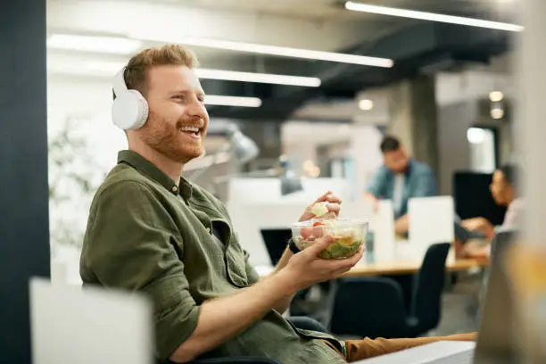 Happy entrepreneur enjoying while eating lunch and listening music over headphones in the office.