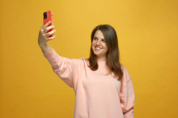 istock Happy girl taking selfie holding phone looking at smartphone using mobile camera smiling having fun on yellow background 1360417929