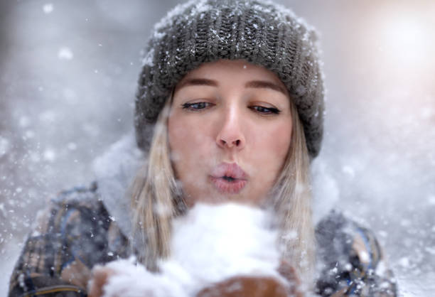 Young woman in a warm hat blows on the snow. Snow is flying straight into the camera. Winter concept