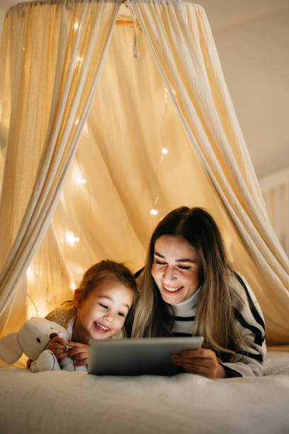 мать и дочь смотрят на свой цифровой планшет вместе, лежа в постели под палаткой. - caucasian child offspring color image стоковые фото и изображения