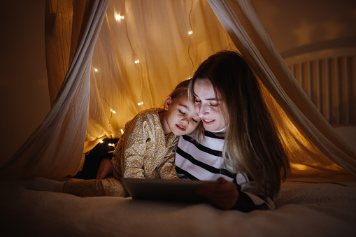 Young mother and cute smiling girl using digital tablet while lying in illuminted hut in kid bedroom. Cheerful woman and lovely daughter on video call under a cozy hut. Lovely little girl with mom watching cartoon on digital tablet in bedroom.