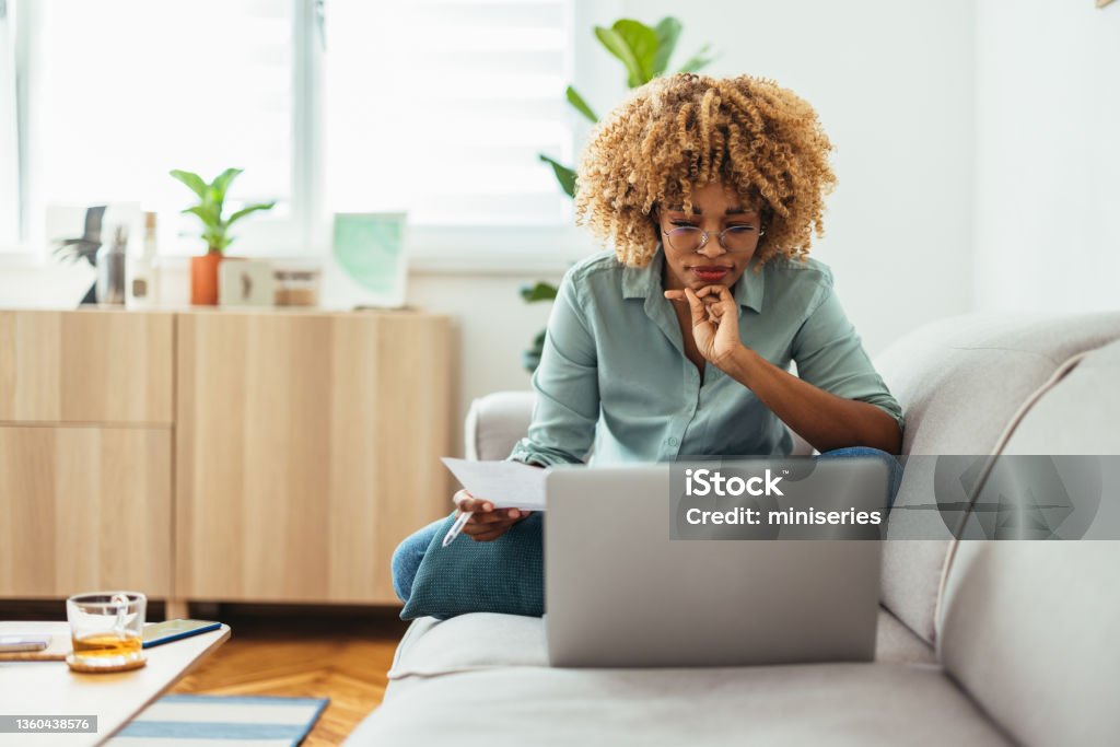 Home Office: An Afro-American Woman Looking at a Laptop Serious Afro-American woman is sitting on a sofa and looking at a laptop while holding a paper in her hand. She is wearing glasses. She might be working from home or studying, or paying a utility bill. She is wearing a green shirt and jeans. Laptop Stock Photo