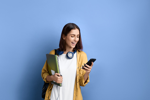 Cheerful student girl is holding mobile phone in hand, looks at screen and laughs, received good exam results, holding books, wearing yellow shirt, white t-shirt, black bag and headphones over neck
