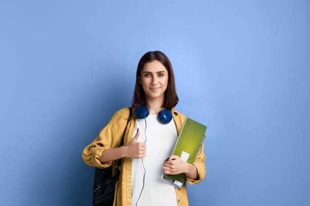 positiv lächelndes studentenmädchen zeigt daumen hoch, mag eine idee, unterstützt initiative, bereit, sich der gruppe anzuschließen, hält bücher, trägt gelbes hemd, weißes t-shirt, schwarze tasche und kopfhörer über dem hals. - thumbs up human thumb human finger brown hair stock-fotos und bilder