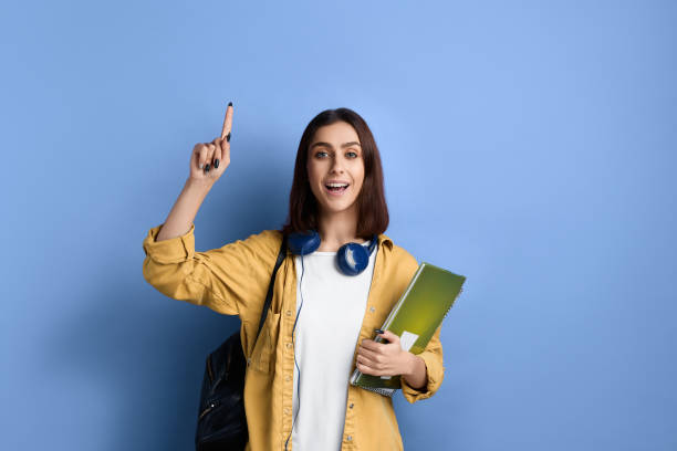 istock Active student girl got insight or idea, is raising index finger up, shows advertisement of school or college, holding books, wearing yellow shirt, white t-shirt, black bag and headphones over neck. 1360442065