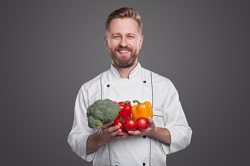 Content bearded male cook in uniform holding raw broccoli with peppers and ripe tomatoes while looking at camera