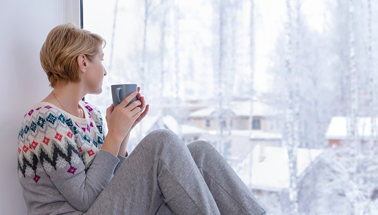 A woman sitting on the windowsill with a cup of tea in winter and looking out the panoramic window