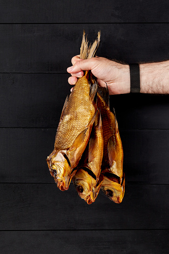 Male hand holding bunch of three appetizing golden smoked breams offering popular fish snacks on black wooden background