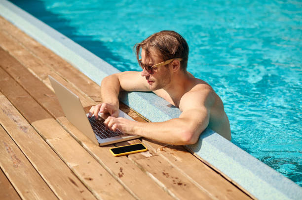 istock Young man at the swimming pool working on a laptop 1360485937
