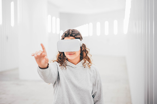 Curly haired young woman in virtual reality glasses moves hands touching online object and looks around standing in white futurisctic tunnel