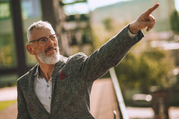 istock Bespectacled gray-haired man showing the direction using a hand gesture 1360498869