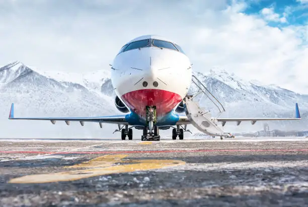 Front view of the modern luxury corporate business jet with open gangway door on the winter airport apron on the background of high scenic mountains