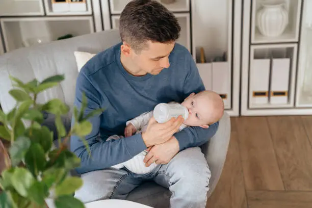Concept of paternity leave instead of maternity one. Happy young dad holding and feeding newborn baby or infant at home while sitting in armchair in living room. Man holding a little baby on his hands