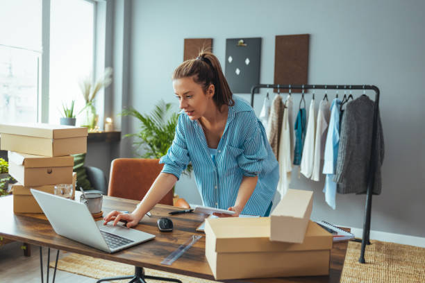 Women, owener of small business packing product in boxes Women, owener of small business packing product in boxes, preparing it for delivery. Working woman at online shop. She wearing casual clothing and checking on laptop address of customer and package information e commerce stock pictures, royalty-free photos & images