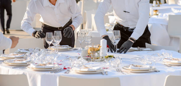 las manos de los camareros con guantes negros protectores organizan una mesa de recepción de fiesta de bodas decorada con flores: platos, tenedores, cuchillos y copas de vino. - restaurant banquet table wedding reception fotografías e imágenes de stock