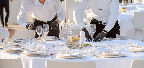 Close up waiters hands in protective black gloves arrange a wedding party reception table decorated with flowers: plates, forks, knives and wine glasses.