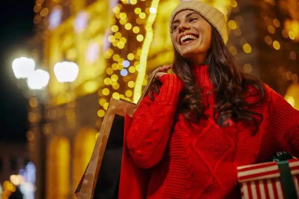 Young adult woman with woolen cap holding wrapped box and shopping bags at Christmas market
