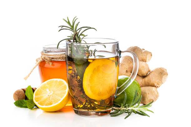 Front view of a mug filled with a hot infused herbs such as rosemary, thyme, mint and lemon surrounded by a ginger root, a honey jar, lime, lemon and herbs. All the objects are at the center of the image isolated on white background.
